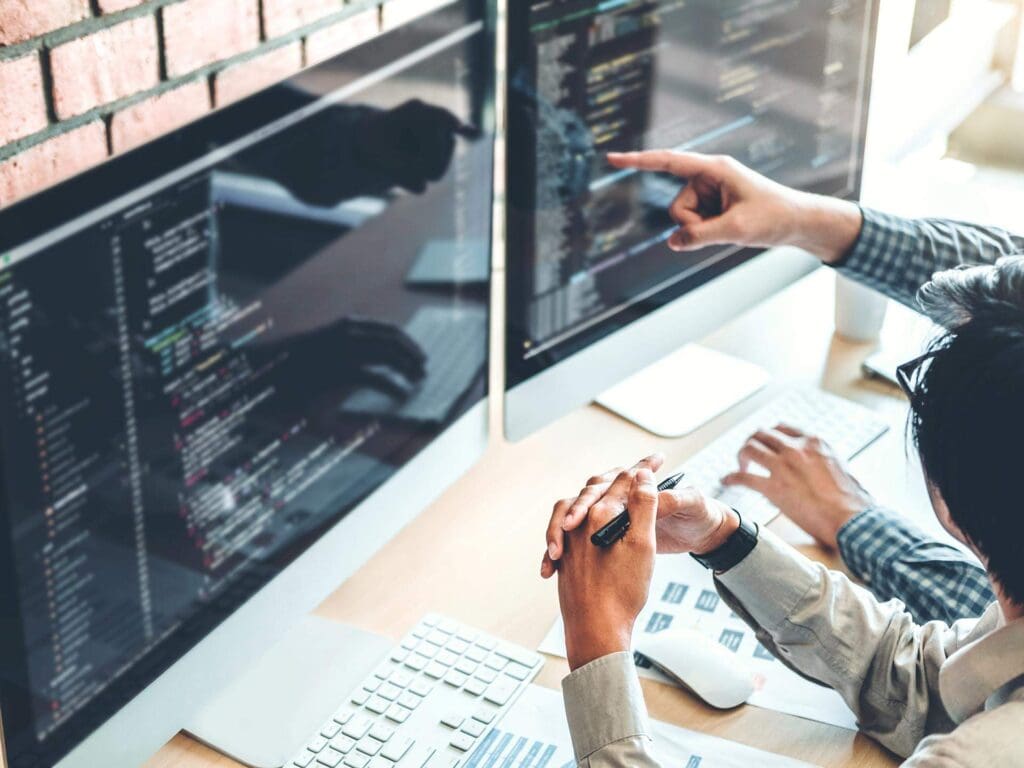 close shot of hands on laptop and a web development code can be seen on three computer screen.