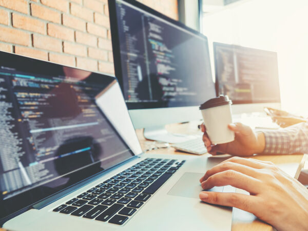 web-developing close shot of hands on laptop and a web development code can be seen on three computer screen.